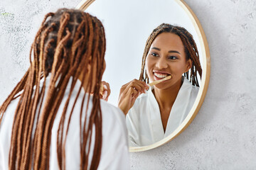 African American woman with afro braids in bath robe brushing teeth in front of a mirror in modern...