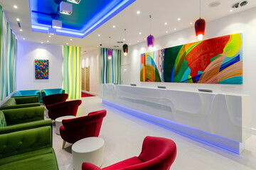 Minimalist white reception counter, green fabric, and red velvet sofa chairs, colorful abstract art on the wall, blue ceiling lights, extra white lighting.