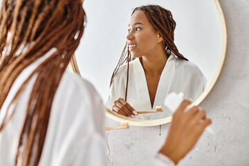 An African American woman in a bathrobe with afro braids brushes her teeth in front of a mirror in...