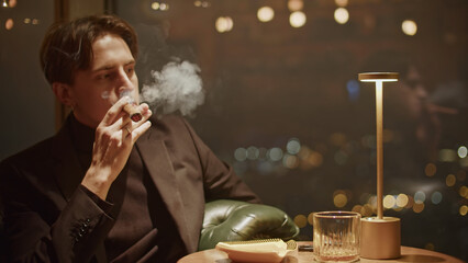 Young man in black clothes smoking cigar at a dark restaurant. Media. Pensive man exhaling smoke.