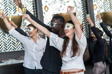 Obraz premium Group of diverse friends toasting with clinking champagne glasses at funny and happy Christmas and new year mask party atmosphere. Holiday celebrate and nightlife lifestyle concept