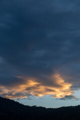 Landscape of thick clouds, orange light hitting the blue sky, silhouettes of trees, forests, and mountains. Area for text in the middle of the image