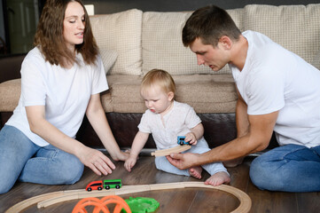 One-year-old child and his young loving parents are sitting on warm floor in cozy nursery, playing...