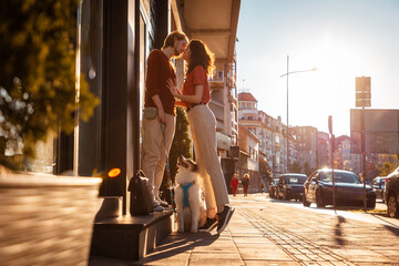 Young couple Caucasian man and woman, are kissing on street. Dog is sitting next to them on leash....