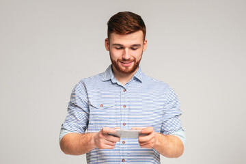 Young guy playing games on his phone and having fun, isolated on grey studio background, free space