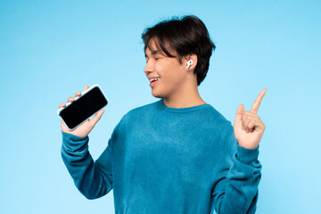 Asian young guy in a blue sweater is happily listening to music on wireless earbuds while holding...