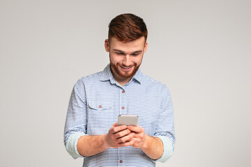 Young man absorbedly text messaging on his cellphone, isolated on grey studio background, free space