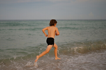 Kid running and splashing in the sea. Summer vacation. Child Running Along the beach. Active little kid running along sea beach during leisure sport activity. Sporty kid running in nature.
