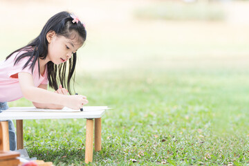 Happy Asian child girl coloring with crayons on paper at park, sitting on grass. Little cute kid with smiling face have fun outdoors activities. Creatives children. Education