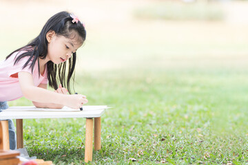 Happy Asian child girl coloring with crayons on paper at park, sitting on grass. Little cute kid...