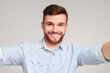 Cheerful handsome bearded redhaired man holding camera and making selfie on beige background,...