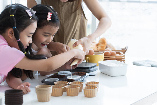 Asian family, mother and two daughters enjoy making cupcakes together at home kitchen. Mom and little child girl squeezing cake batter into cupcake paper cup, prepare for bake, smiling. Happy holiday