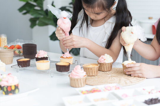 Asian little cute child girl enjoy making cupcakes with friend at home kitchen, squeezing cream from piping bag to decorate homemade cupcake, smiling with holiday activities. Selective focus on hands