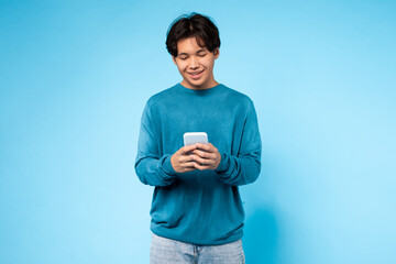 Asian guy wearing a blue sweater is sitting or standing and looking intently at a cell phone in his...