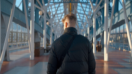 Rear view of man walking in station building. Action. Man walks along sunny corridor of stylish...
