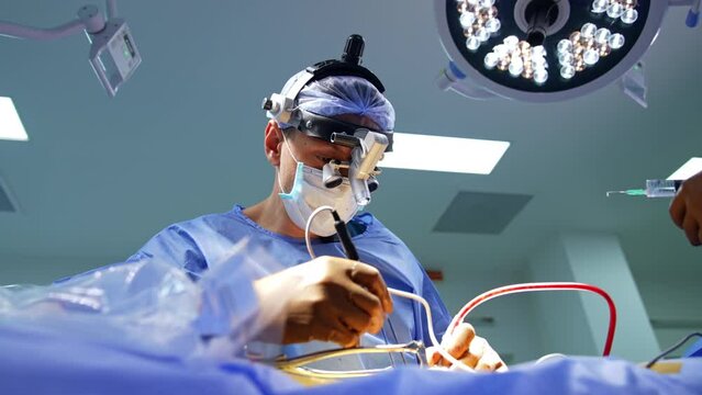 Male neurosurgeon takes the electric tools device. Doctor in surgical loupes carries out the operation assisted by female nurse. Low angle view.