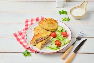 Potato pancakes stuffed with minced meat on a ceramic plate on a light wooden background. Served with fresh cucumber and cherry tomatoes. Belarusian cuisine.
