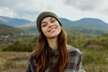 Smiling young woman in plaid shirt and beanie standing in front of majestic mountains enjoying a...