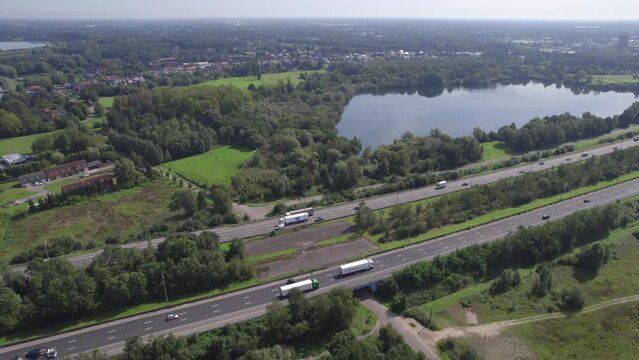 Aerial footage showcasing the E19 highway running between Brussels and Antwerp as seen from Mechelen. The video captures a stretch of the highway bordered by lush greenery, a tranquil lake, and