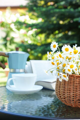 Bouquet of flowers in basket with book, cup and coffee pot. Bouquet of chamomile flowers. Spring, summer holidays, Easter, mother's day. Side view. Space for text.