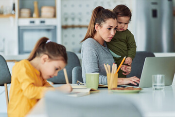 A focused single mother using a laptop and working from home while taking care of her kids