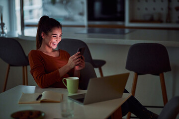A happy young adult woman scrolling through her phone while working late