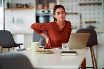 A distraught adult female freelancer rubbing her sore back while working from home on a laptop