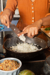 Close-up of Hispanic woman's hand in kitchen pouring chopped onion into frying pan with oil