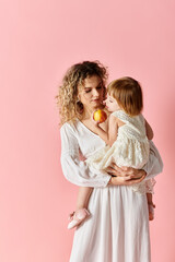 A woman holding a child while eating an apple on a pink background.