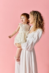 A mother in a white dress lovingly holds her baby girl in a white dress on a soft pink background.