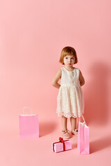 Adorable girl in white dress with gift bags against pink backdrop.