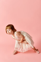A little girl in a white dress stands charmingly against a pink backdrop.
