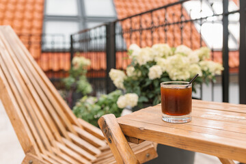 A glass of iced coffee on a wooden table next to a wooden chaise longue on the terrace.