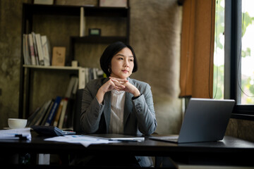 A woman in a gray jacket and white shirt sits at a desk with a laptop and a cup