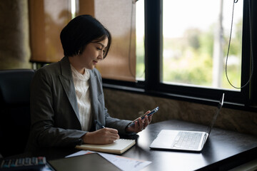 A woman in a business suit is sitting at a desk with a laptop and a cell phone
