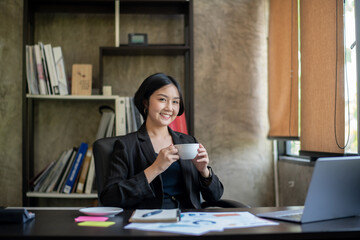 A woman in a black suit is sitting at a desk with a laptop and a cup of coffee
