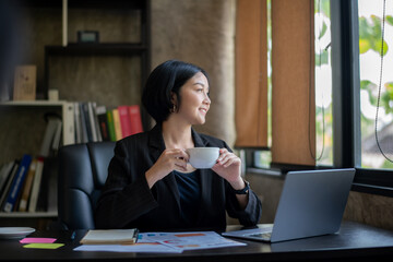 A woman in a black suit is sitting at a desk with a laptop and a cup of coffee