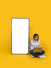 Mobile application advertising design,  full body size woman sit use work laptop near big huge empty blank screen mobile phone mock up. Yellow studio background. Technology concept idea image.