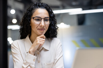 A professional businesswoman with curly hair and glasses works confidently and focused on a laptop...