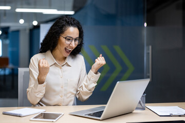 Curlyhaired woman with glasses celebrates work success in a modern office, raising her fists...