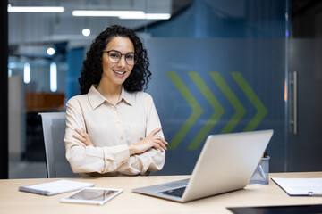 Confident businesswoman with curly hair smiling and sitting at her desk in a modern office, working...