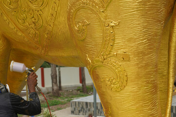 An artist spray paints an elephant statue with gold paint.