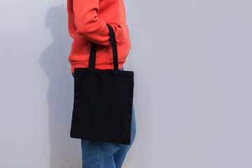 Woman in red hoodie holding tote canvas blank eco bag on street grey minimal wall background. Female consumer hold black textile shopper. template or place for your design, text, logo