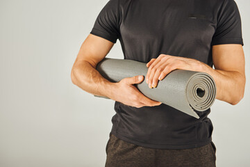 Young sportsman in active wear holds a yoga mat in a studio with a grey background.