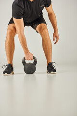 A young sportsman in active wear is squatting with a kettlebell in a studio with a grey background.