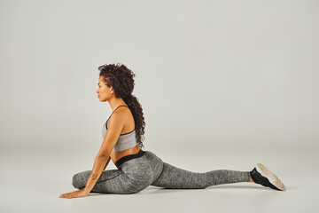 A young African American woman in active wear gracefully stretches in a squat position on a grey...