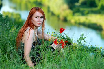 A girl with wild poppies enjoys the view from a hill near the river. Joy, happiness, serenity. Wild poppies by the river.
