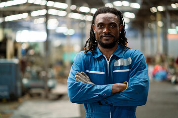 A man in a blue uniform stands in a factory with his arms crossed. He has a serious expression on...