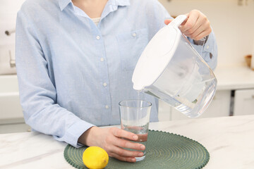 Woman pouring water from filter jug into glass at white marble table in kitchen, closeup