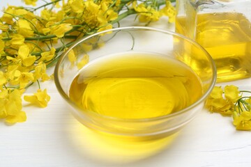 Rapeseed oil in bowl and beautiful yellow flowers on white wooden table, closeup